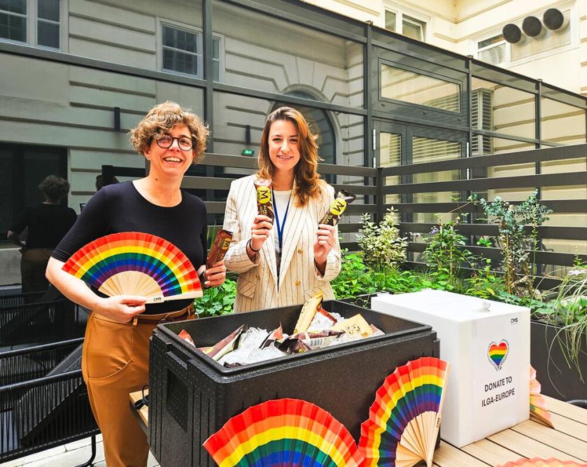 Fundraiser in the courtyard of CMS Vienna, an employee holds a rainbow fan, the employee next to her holds a Magnum ice cream in front of a donation box with a rainbow heart and the inscription &ldquo;Donate to ILGA-Europe.&rdquo;