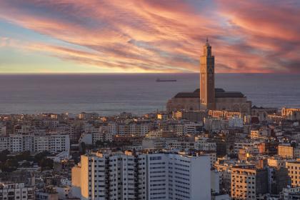 Vue panoramique de la ville de Casablanca au coucher du soleil, avec au premier plan des immeubles blancs et beige, et en arrière-plan la mosquée Hassan II dominant le littoral. Le ciel est rempli de nuages aux teintes rosées et orangées, reflétant la lumière du crépuscule, tandis que l’océan Atlantique s’étend à l’horizon avec un navire visible au loin.