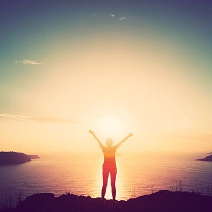 woman with hands up standing on cliff over sea and islands at sunset