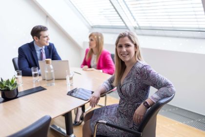 Drei Personen im Besprechungszimmer des hellen CMS Büros in Wien, zwei Personen sind im Gespräch und eine lächelnde Frau mit Tablet am Tisch blickt in die Kamera